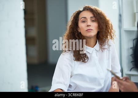 Attraktive junge braune Frau mit langen lockigen Haaren, die in reinweißem, geknöpftem Hemd steht. Sie saß auf der Bürocouch und schaute nachdenklich in die Ferne Stockfoto