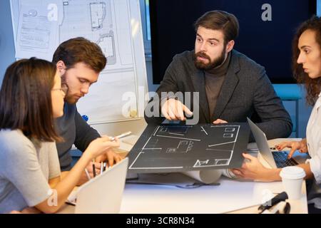 Junge männliche und weibliche Spezialisten sitzen am Schreibtisch, mit Hausentwurf in der Mitte. Stilvoller bärtiger Mann, der auf einen Plan zeigt und Ideen zur Innenarchitektur mit Kollegen teilt Stockfoto
