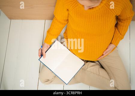 Overhead-Aufnahme einer zukünftigen Mutter, die eine wichtige To-do-Liste für die Mutterschaftszeit schreibt. Detaillierte Organisation der Spitaltasche und Geburtsvorbereitungsnotizen. Stockfoto