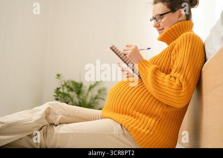 Heitere schwangere Frau schreibt wichtige Liefererinnerungen zu Hause im Notizbuch. Durchdachte Vorbereitung auf die Grundlagen der Entbindungs- und Neugeborenenpflege Stockfoto