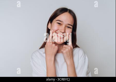 Lächelnde junge asiatische Frau, die ihre Freude mit verspielten Gesten in einer minimalistischen Umgebung zum Ausdruck bringt Stockfoto