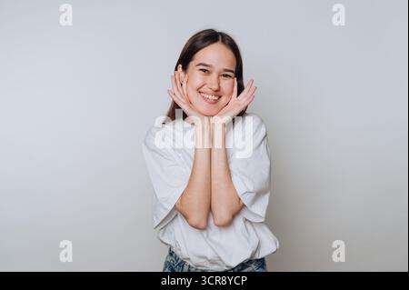 Junge Frau, die bei Tageslicht in einem Studio lächelt, mit Händen auf den Wangen Stockfoto