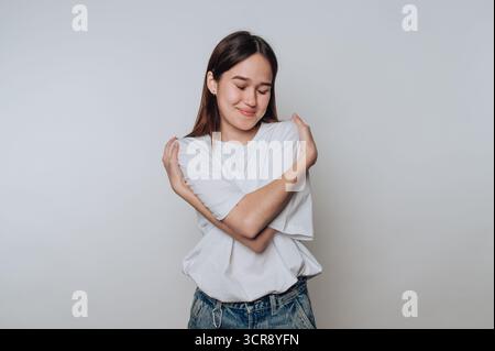 Lächelnde junge asiatische Frau umarmt sich in einem gemütlichen Innenraum während eines ruhigen Moments Stockfoto