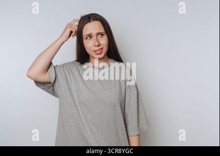 Junge Frau, die Verwirrung ausdrückt, während sie in einer ungezwungenen Innenumgebung mit neutralem Hintergrund denkt Stockfoto
