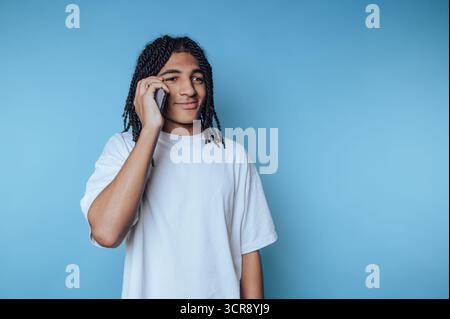 Junge Person, die am Telefon vor hellblauem Hintergrund mit einem entspannten Ausdruck in einer ungezwungenen Umgebung spricht Stockfoto