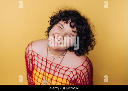 Lächelnde junge Frau in farbenfrohen Outfit posiert vor hellgelbem Hintergrund, während sie fröhlichen Ausdruck annimmt Stockfoto