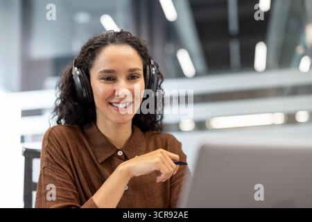 Frau, die Kopfhörer trägt, lächelt während eines Online-Meetings in einem modernen Büro auf den Laptop-Bildschirm, nimmt an einer virtuellen Konferenz Teil, professionell und selbstbewusst in der Remote-Arbeit-Umgebung Stockfoto