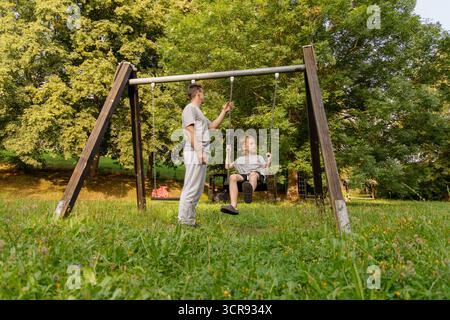 Ein Vater schiebt seinen kleinen Sohn auf eine Schaukel auf einem Spielplatz in einer grünen Gegend. Sie teilen einen freudigen Moment zusammen und zeigen die Verbundenheit von Stockfoto