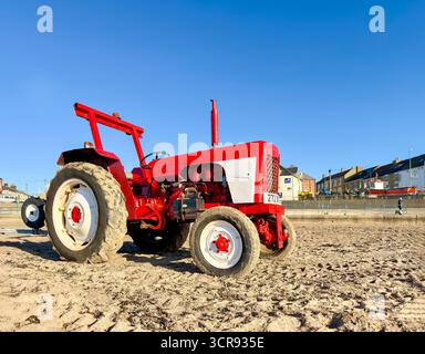 Roter David Brown Traktor, der an einem Sandstrand mit Gebäuden an der Küste und einem hellblauen Himmel im Hintergrund geparkt wurde Stockfoto