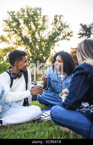Verschiedene Gruppen von Universitätsstudenten sitzen auf dem Campus, plaudern, Snacks teilen und lachen während einer Pause, entspannt und gesellig in einem hellen Ambiente Stockfoto