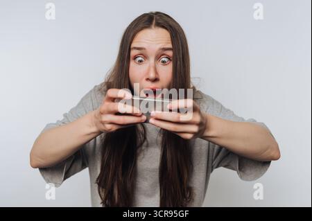 Die junge Frau drückt ihre Begeisterung aus, während sie auf ihrem Smartphone auf weißem Hintergrund ein mobiles Spiel spielt Stockfoto