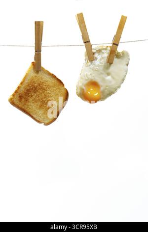 Geröstete Brotscheibe und Spiegelei, die an einer Wäscheleine auf weißem Hintergrund hängen, stellen eine einfache und schnelle Frühstückslösung dar Stockfoto