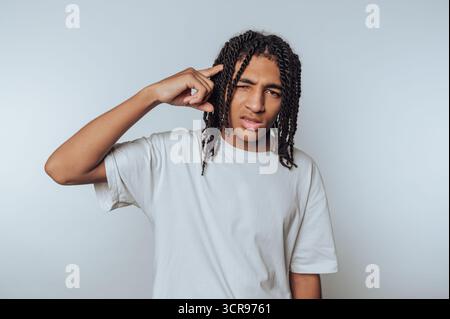 Junger Mann mit geflochtenen Haaren zeigt einen nachdenklichen Ausdruck, während er auf seinen Tempel auf weißem Hintergrund zeigt Stockfoto