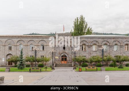 Blick auf das alte Steingebäude mit Bogenfenstern und einer kleinen Fahne, die oben flattert, umgeben von grünen Bäumen und Bänken, Goris, Provinz Syunik, Armenien. Stockfoto