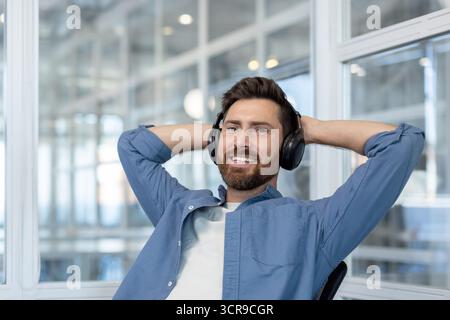 Glücklicher bärtiger Mann, der sich in einem Bürostuhl mit Händen hinter dem Kopf zurücklehnt und eine Pause von der Arbeit genießt, während er Audiosignale hört, um sich zu entspannen und sich ruhig zu fühlen Stockfoto