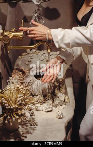 Eine junge Frau mit modischen Perlen wäscht sich die Hände in einem einzigartigen Waschtisch mit Muscheln. Die Szene fängt einen Moment des Elegans ein Stockfoto