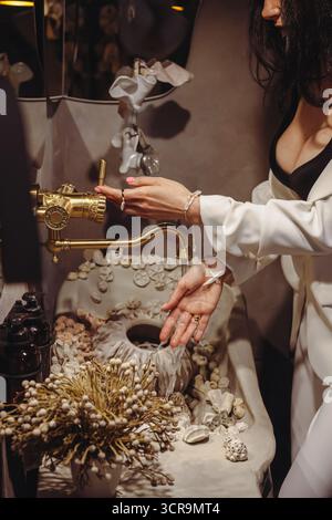 Eine junge Frau, die stilvolle Perlenperlen in verschiedenen Formen trägt, reinigt ihre Hände in einem wunderschön gestalteten Waschtisch mit Muscheln und schafft so einen Ser Stockfoto