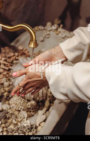 Eine junge Frau mit stilvollen Perlen wäscht sich die Hände in einem dekorativen Waschtisch mit Muscheln. Die Umgebung ist elegant und einladend, Show Stockfoto