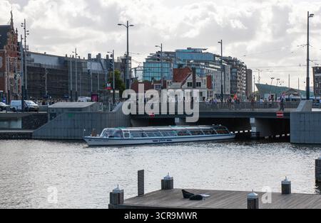 Amsterdam, Niederlande - 5. August 2025: Ein Boot gleitet unter einer Brücke, während die Menschen entlang der nahe gelegenen Uferpromenade spazieren. Stockfoto
