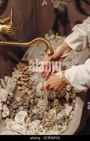 Eine junge Frau mit modischen Perlen in verschiedenen Formen wäscht sich die Hände an einem wunderschön verzierten Waschtisch, gefüllt mit Muscheln. Sie genießt einen Stockfoto