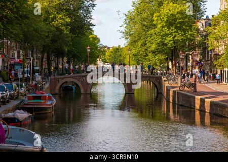 Amsterdam, Niederlande - 5. August 2025: Menschen schlendern entlang des Kanals, während Boote unter einer malerischen Brücke in Amsterdam schweben. Stockfoto