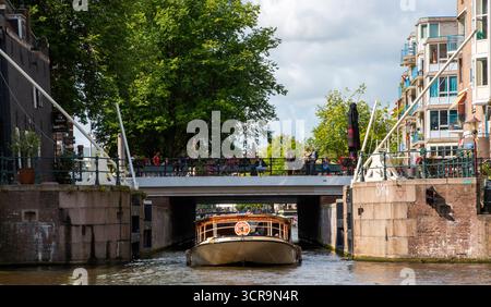 Amsterdam, Niederlande - 5. August 2025: Ein Ausflugsboot gleitet sanft unter einer Brücke auf einem Kanal, während die Menschen die Aussicht von oben genießen. Stockfoto