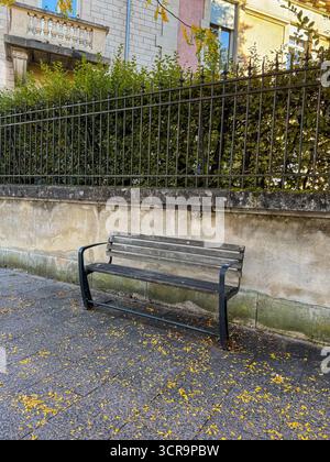 Leere Holzbank, umgeben von gefallenen gelben Blättern auf einem Kopfsteinpflaster-Gehweg, mit einer alten Steinmauer und einem schmiedeeisernen Zaun, die an die autulente Ruhe erinnern Stockfoto