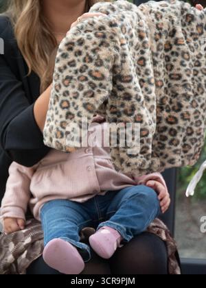 Rom, Italien - 11. September 2025 entzückendes Kleinkind mit kuscheliger Jacke mit Leopardenmuster und Denim-Jeans, das sich mit einem liebevollen Elternteil Outdoo warm kuschelt Stockfoto