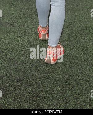 Nahaufnahme von weiblichen Beinen in roten Turnschuhen und blauen Jeans, die auf dem grünen Sportplatz spazieren. Konzept von aktivem Lifestyle, Fitness, Casual Fashion und Healt Stockfoto