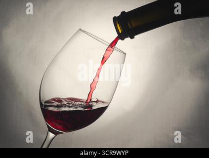 Rotwein wird in ein Stängelglas gegossen. Eine Flasche Rotwein gießt einen Strom Wein in ein Weinglas, vor hellem Hintergrund. Weinblase Stockfoto