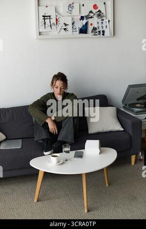 Eine junge Frau sitzt leise auf einer Couch, in Gedanken verloren, umgeben von einer beruhigenden Umgebung. Stockfoto