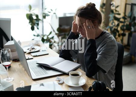 Eine junge Frau spürt das Gewicht der Depression, während sie zu Hause an einem überladenen Schreibtisch sitzt. Stockfoto