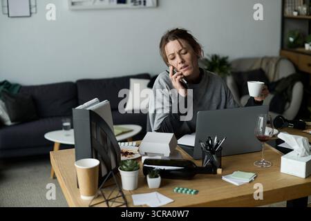 Eine junge Frau sitzt an ihrem Schreibtisch und fühlt sich überwältigt, während sie am Telefon spricht, während sie eine Tasse hält. Stockfoto