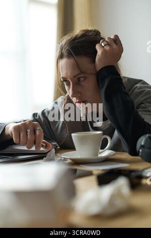 Eine Frau sitzt allein an einem Tisch, sichtbar beunruhigt, während sie eine Zigarette hält und still starrt. Stockfoto