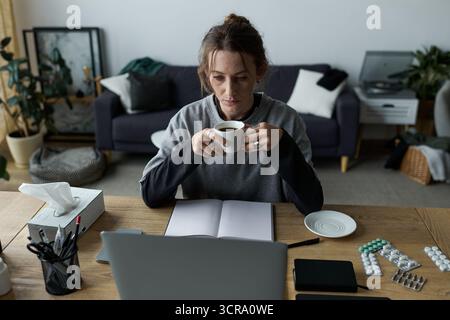 In einem gemütlichen Wohnzimmer sieht eine junge Frau nachdenklich aus, während sie allein an ihrem Schreibtisch Kaffee schlürft. Stockfoto