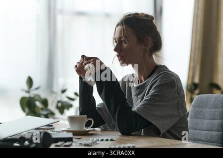 Eine junge Frau sitzt allein zu Hause, verloren in ihren Gedanken mit einem Becher in der Hand. Stockfoto
