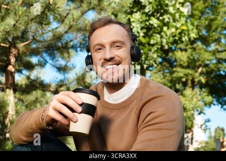 Junger männlicher Pädagoge entspannt sich in einem grünen Park, schlürft Kaffee und hört Musik mit Kopfhörern. Stockfoto