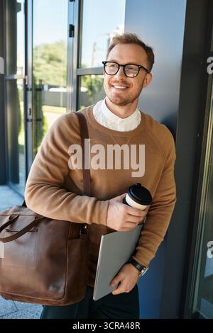 Ein junger Mann, lässig gekleidet, hält eine Kaffeetasse und einen Laptop, während er draußen posiert. Stockfoto