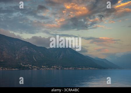 Monduntergang vor Sonnenaufgang über Kotor Bay, Montenegro, mit nebeligen Bergen, weichen Wolken und rosa-blauem Himmel, der sich vor Sonnenaufgang in stillen Gewässern spiegelt Stockfoto
