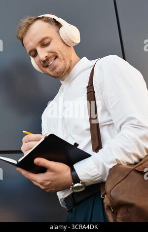 Ein junger Universitätslehrer schreibt Notizen in ein Notizbuch, trägt Kopfhörer und lächelt. Stockfoto