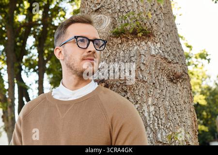 Ein junger Mann, ein Universitätslehrer, posiert neben einem großen Baum, während er in Gedanken verloren ist. Stockfoto