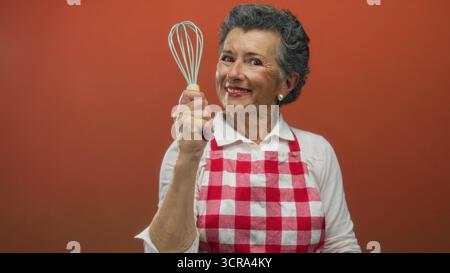 Eine ältere Frau mit grauem Haar in einer roten karierten Schürze hält fröhlich einen Schneebesen vor einem orangefarbenen Hintergrund und zeigt ihre Leidenschaft für Kochen und Küche Stockfoto