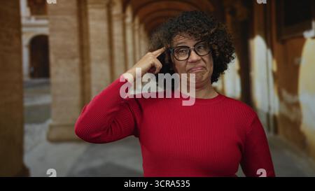 Hispanische Frau im mittleren Alter in rotem Pullover zeigt mit dem Finger auf den Tempel auf der Kopfsteinpflasterstraße; Frustration. Stockfoto