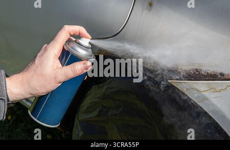 Nahaufnahme der Hand eines Mannes, der eine rostige Autooberfläche mit einer Sprühdose mit weißer Sprühfarbe bedeckt. Stockfoto