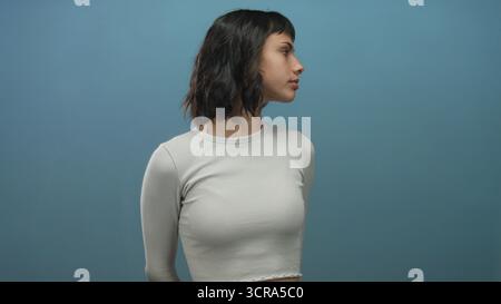 Die junge hispanische Frau dreht sich Kopf an Seite und zeigt ein Gesichter mit weißem geripptem Oberteil in blauem Studio; Besinnung. Stockfoto