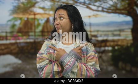 Frau mit gekreuzten Armen und Fingern, die in beide Richtungen auf Straßengeländer unter Bäumen zeigen, Brille und gemusterte Jacke tragen; Ungewissheit. Stockfoto