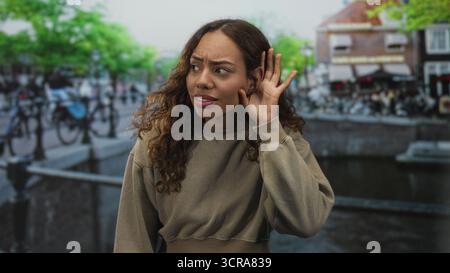 Frau, die Hand an Ohr auf einem Straßenkanal in amsterdam schröpft und beigefarbenes Sweatshirt an Brücke und Fahrrädern trägt; Neugier hört zu. Stockfoto