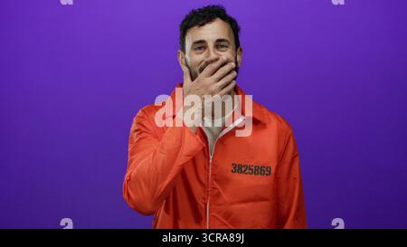 Hispanischer Mann im orangefarbenen Gefängnis-Jumpsuit, der Mund mit der Hand vor einem lebhaften lila Hintergrund bedeckt und einen überraschten Ausdruck in einem Stockbild zeigt Stockfoto