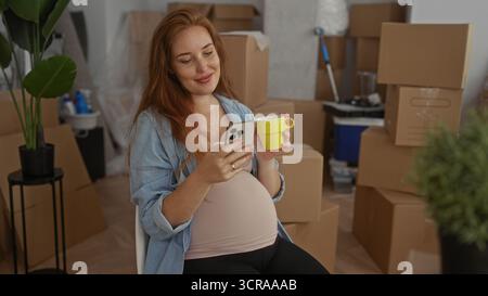 Schwangere Frau hält hellgelben Becher und Smartphone gegen sichtbaren Bauch im Gebäude; Mutterschaft Hoffnung. Stockfoto