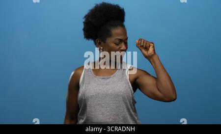 Frau beugt Muskeln in lässiger Kleidung vor isoliertem blauem Hintergrund und zeigt Empowerment und Kraft. Stockfoto